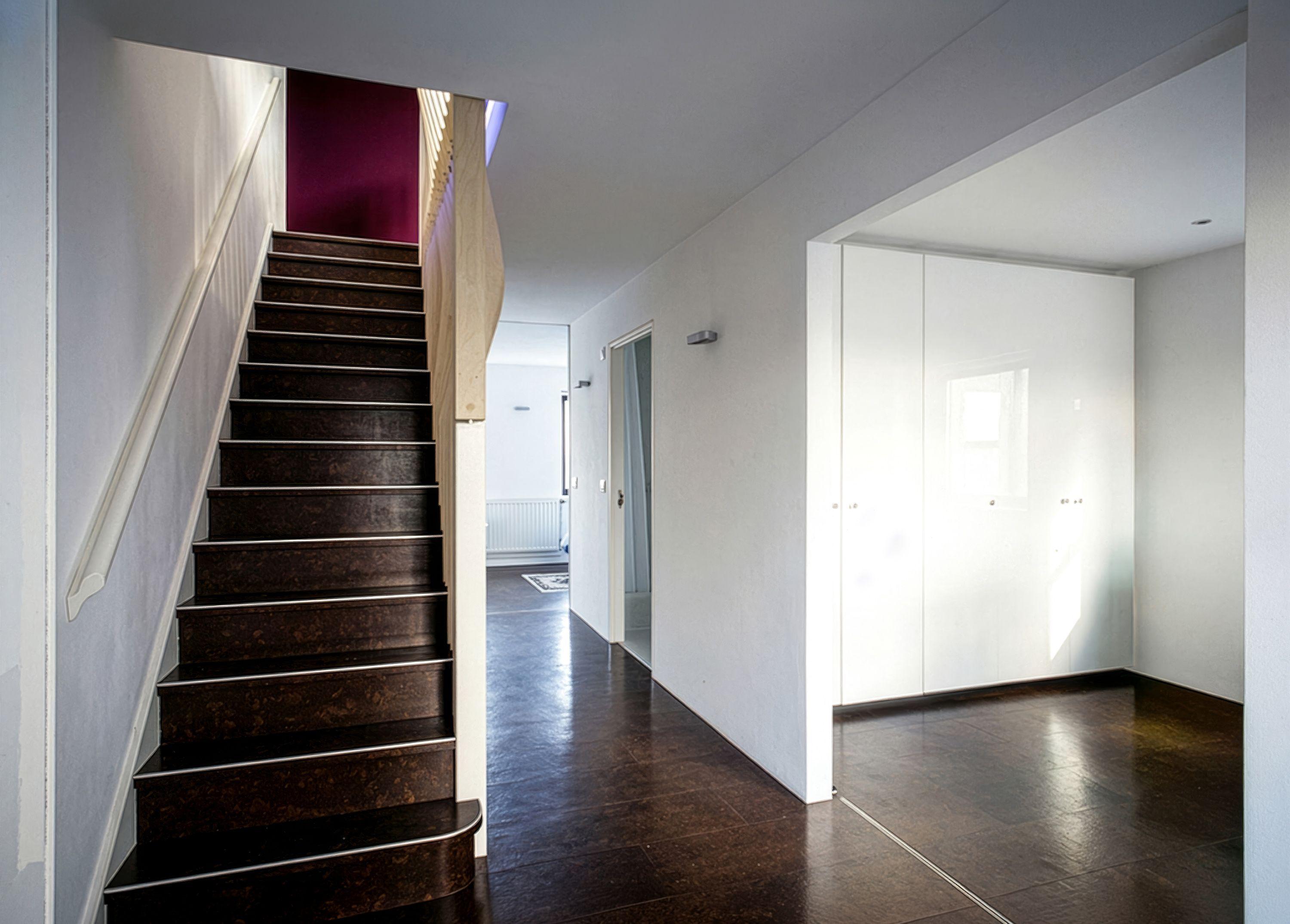 An apartment hallway with a dark cork-brown stair rising past a timber-fin balustrade toward a plum-fabric wall, white joinery cupboards to one side