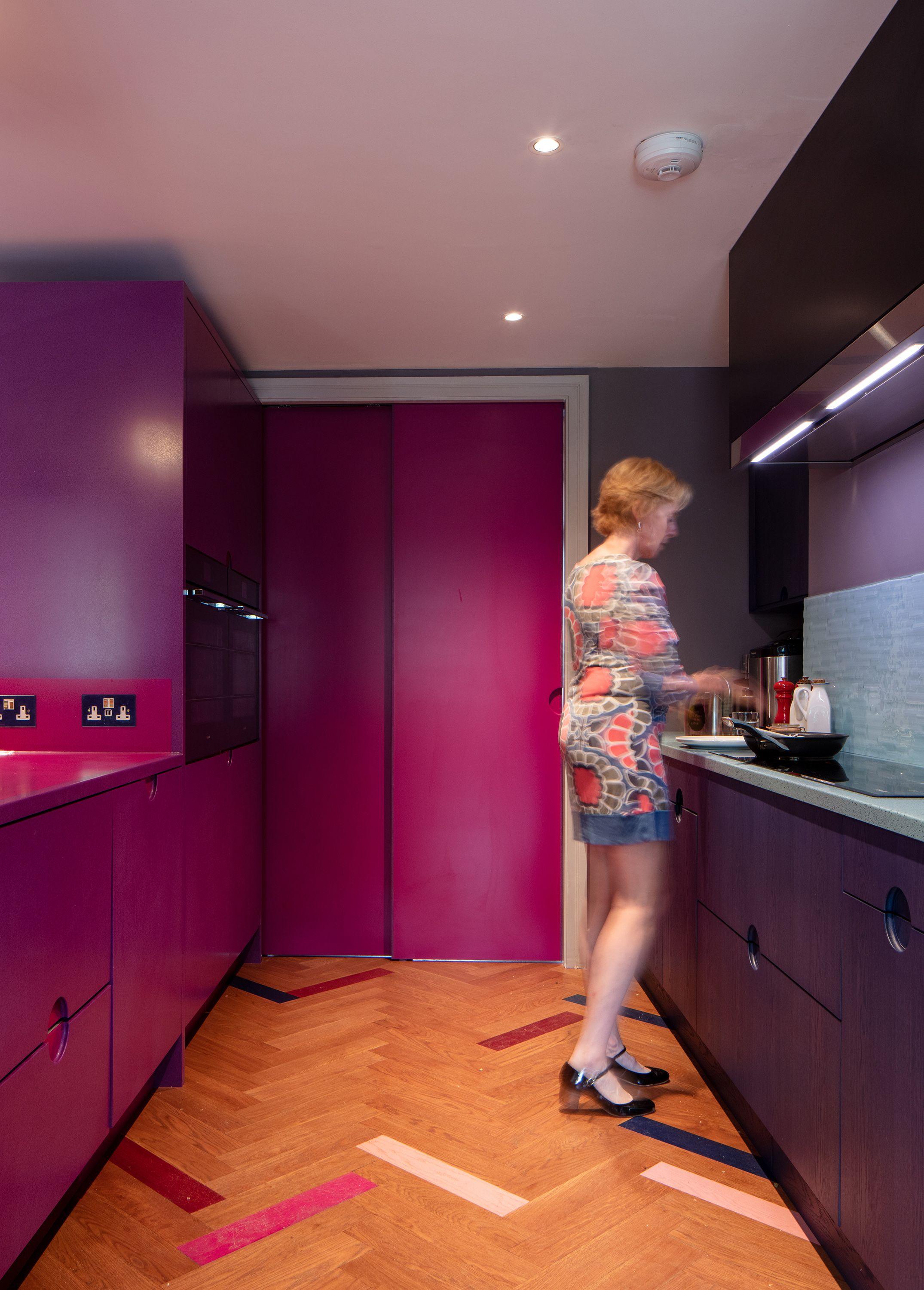 Kitchen at Puzzle Box with a magenta sliding door at its end, herringbone floor inset with coloured blocks, a figure at the counter.