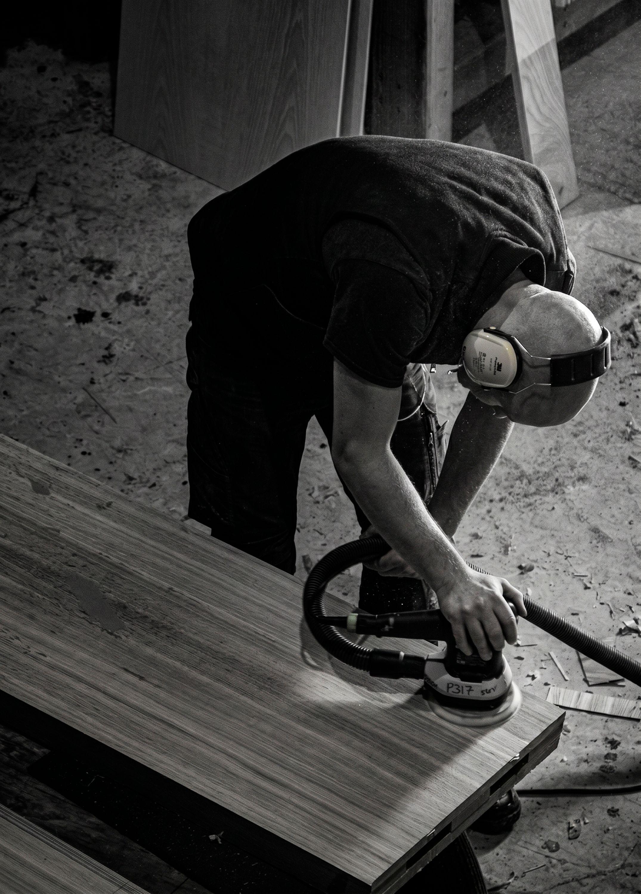 A joiner sanding a plywood panel for the Hedgerow at Regent's Place with an orbital sander connected to dust extraction in the workshop.