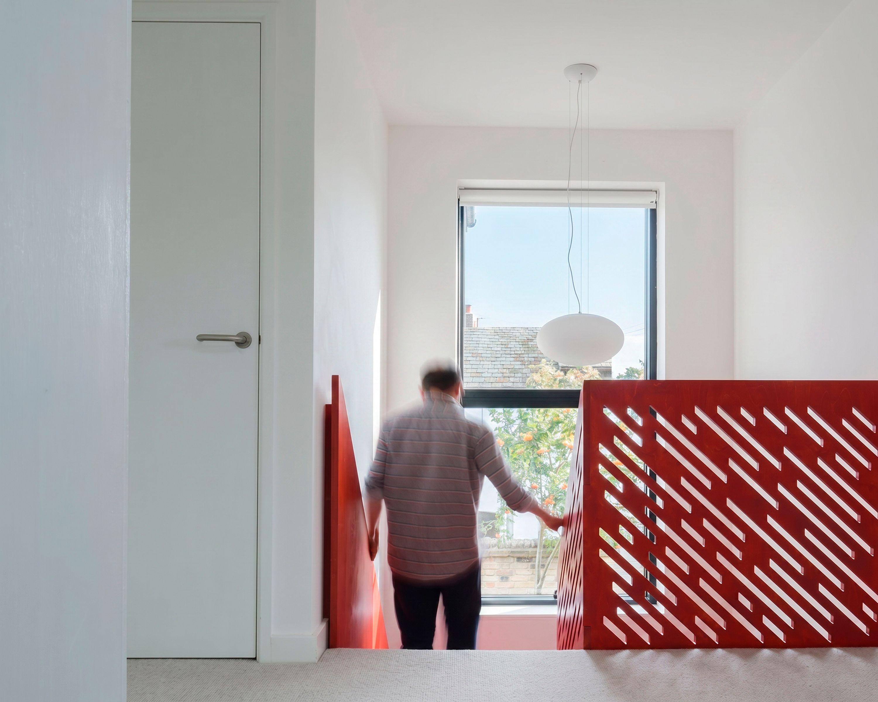 Upper landing of Cross Cut House with a red plywood balustrade cut in a diagonal slot pattern, a figure heading down the stair toward a garden window.