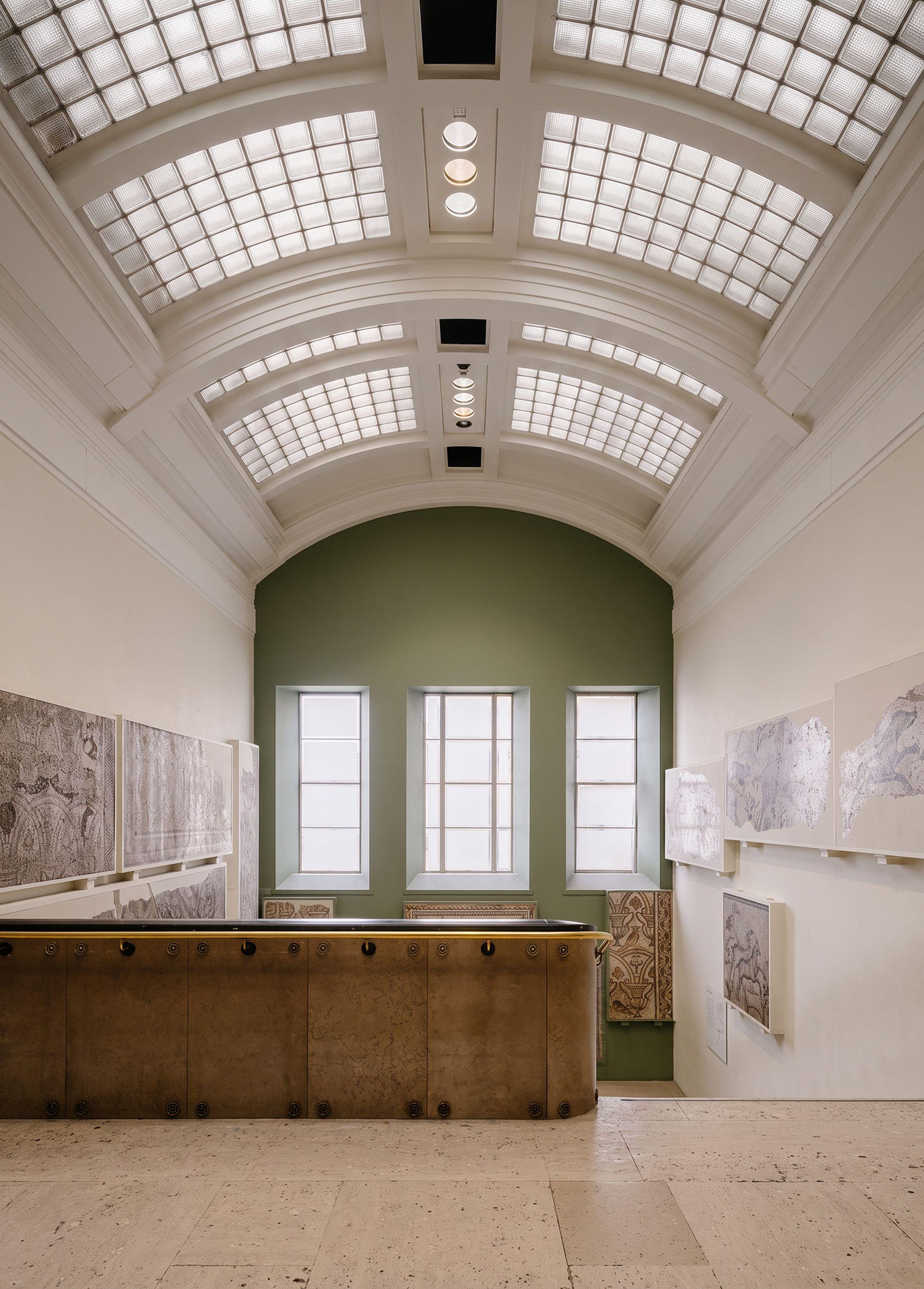 Stair gallery in the British Museum's North Wing, Assyrian reliefs lining the walls beneath a barrel-vaulted ceiling with replacement double-glazed rooflights and a deep green apse wall.