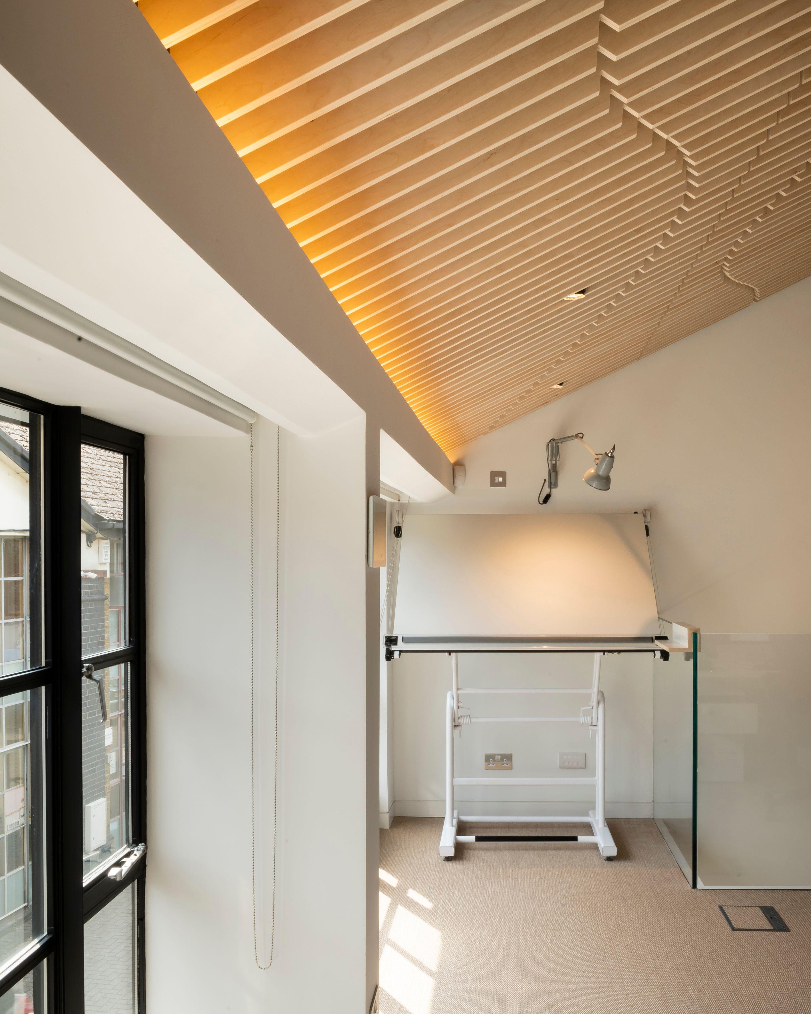 Daylit studio corner at the Fluid Workspace, an angled drafting table below the battened timber roof, tall black-framed windows alongside.