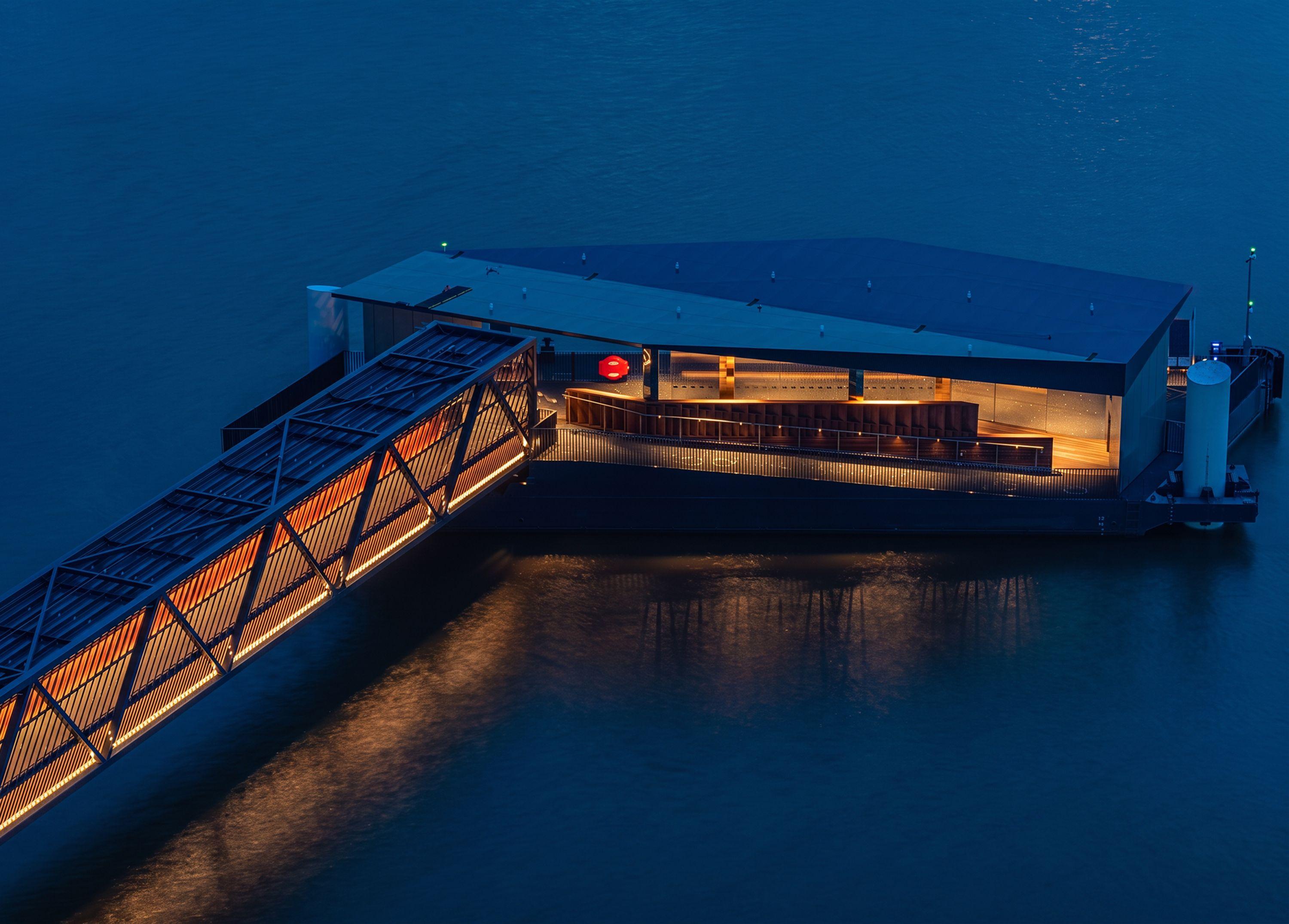 Royal Wharf Pier at night seen from above, the lit floating walkway leading to the sheltered waiting area and pontoon, its folded roof reading dark against the Thames.