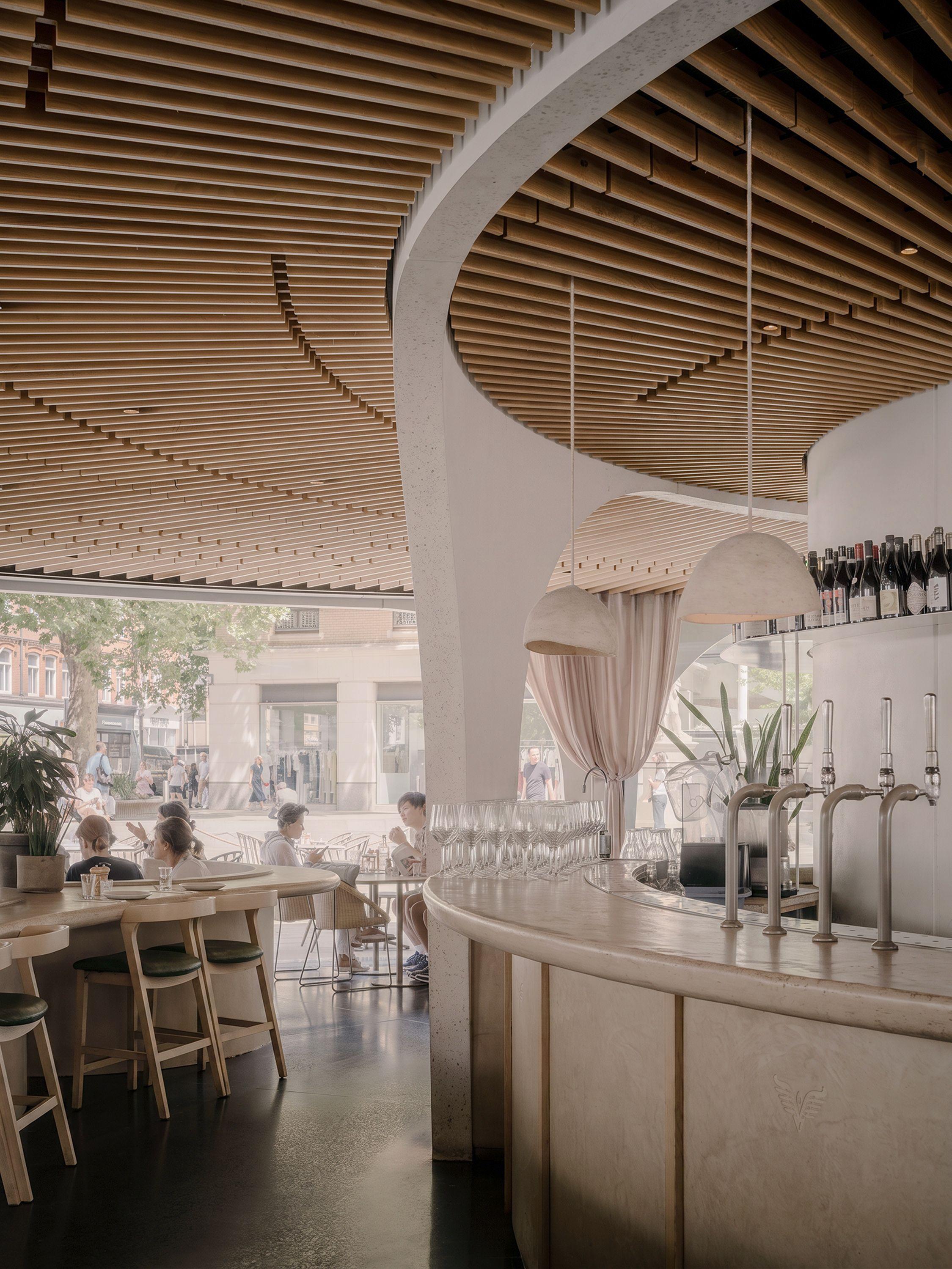 Interior of Vardo, Duke of York Square, London, its curved service core rising to an acoustic ceiling of arranged ashwood slats, with a stone-clad bar and diners at the counter.