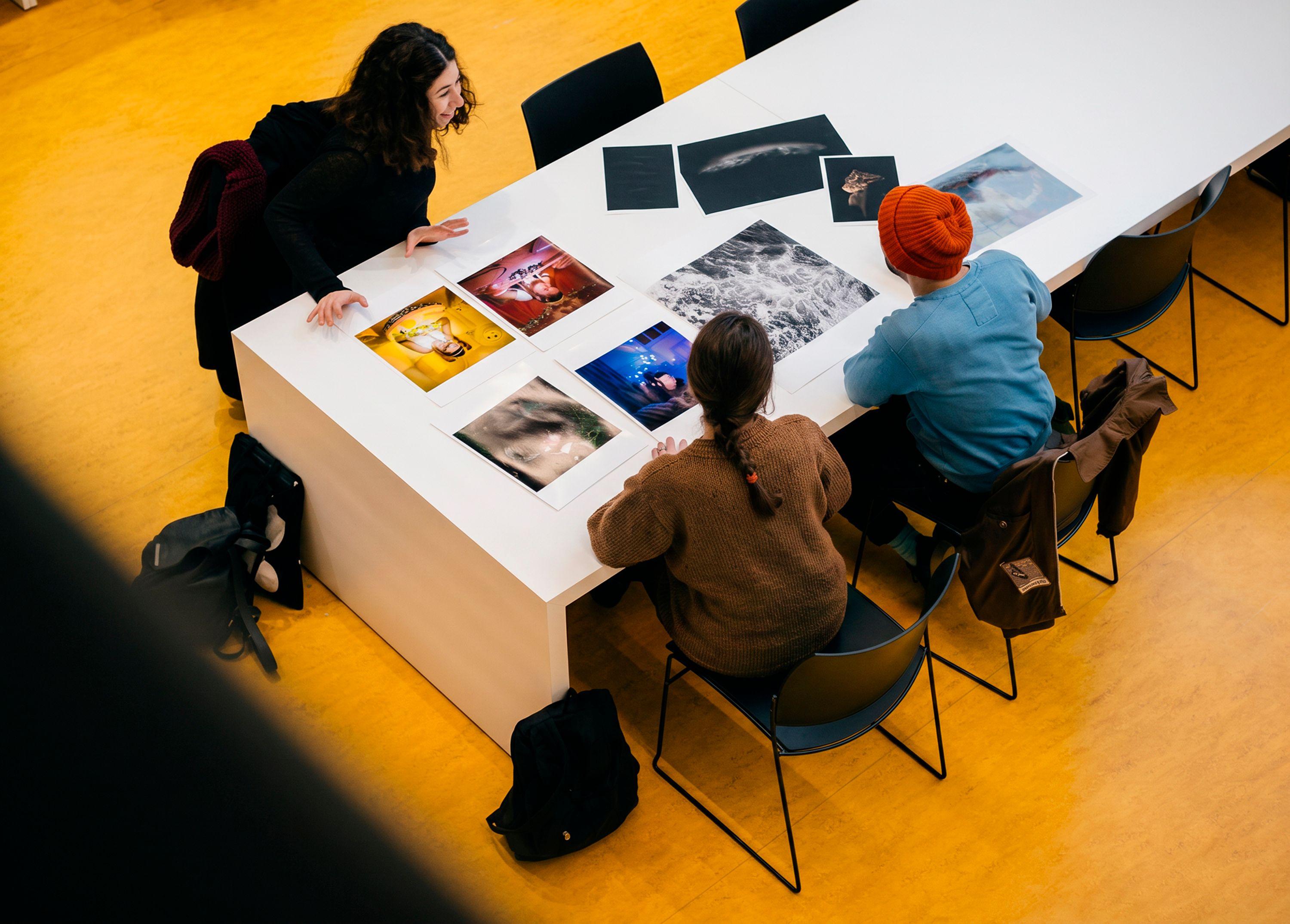 : Overhead view of three students editing a selection of prints on a table in Brighton, the yellow flooring marking the production spaces below.