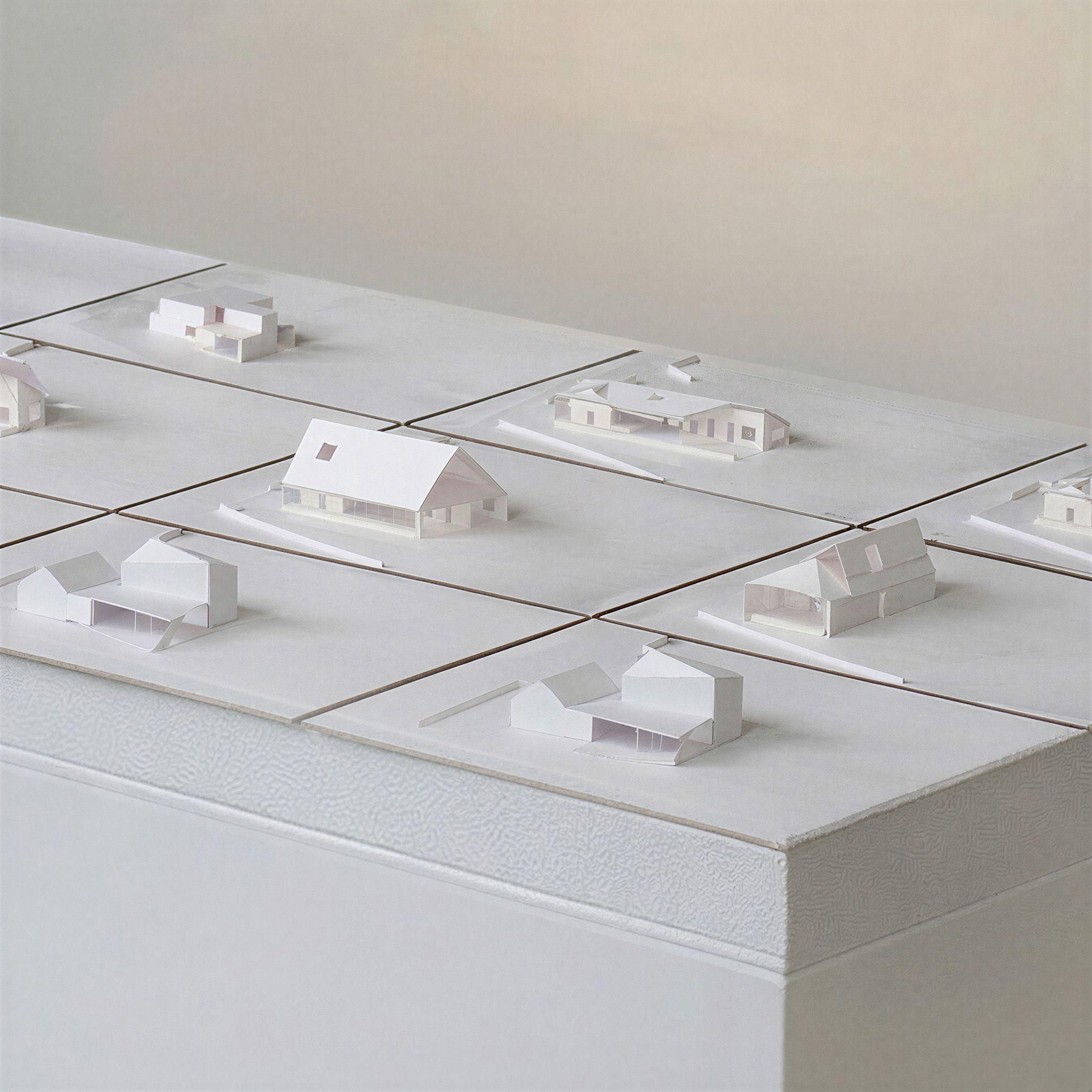 Close view of the Keeping House study models, each white card model on its own base testing a different pairing of the gabled cottage with the volume behind.