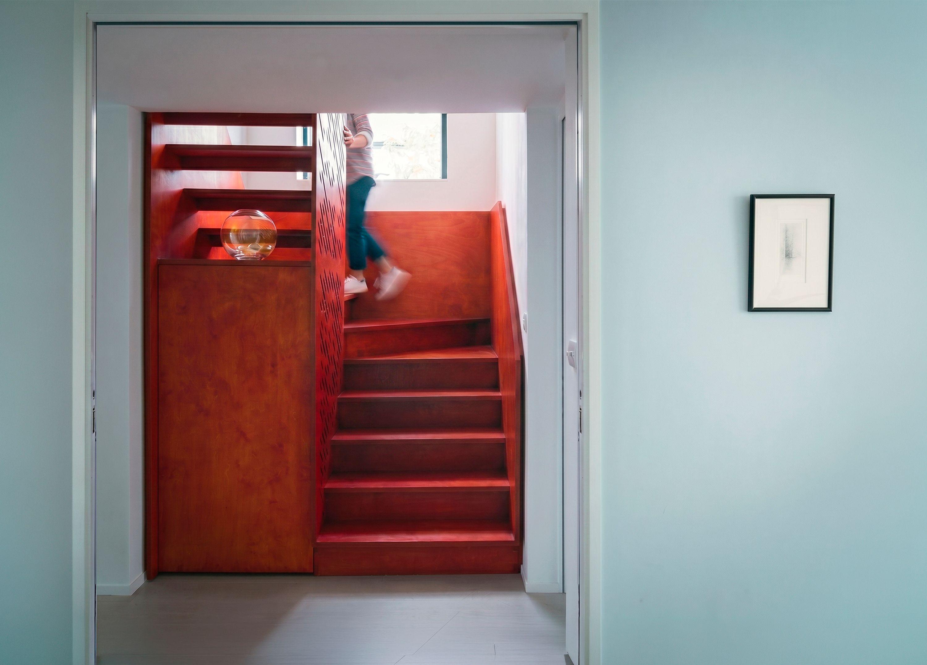 The red plywood stair of Cross Cut House framed by a doorway, a glazed bowl on the built-in cabinet and a small framed drawing on the pale blue wall alongside.