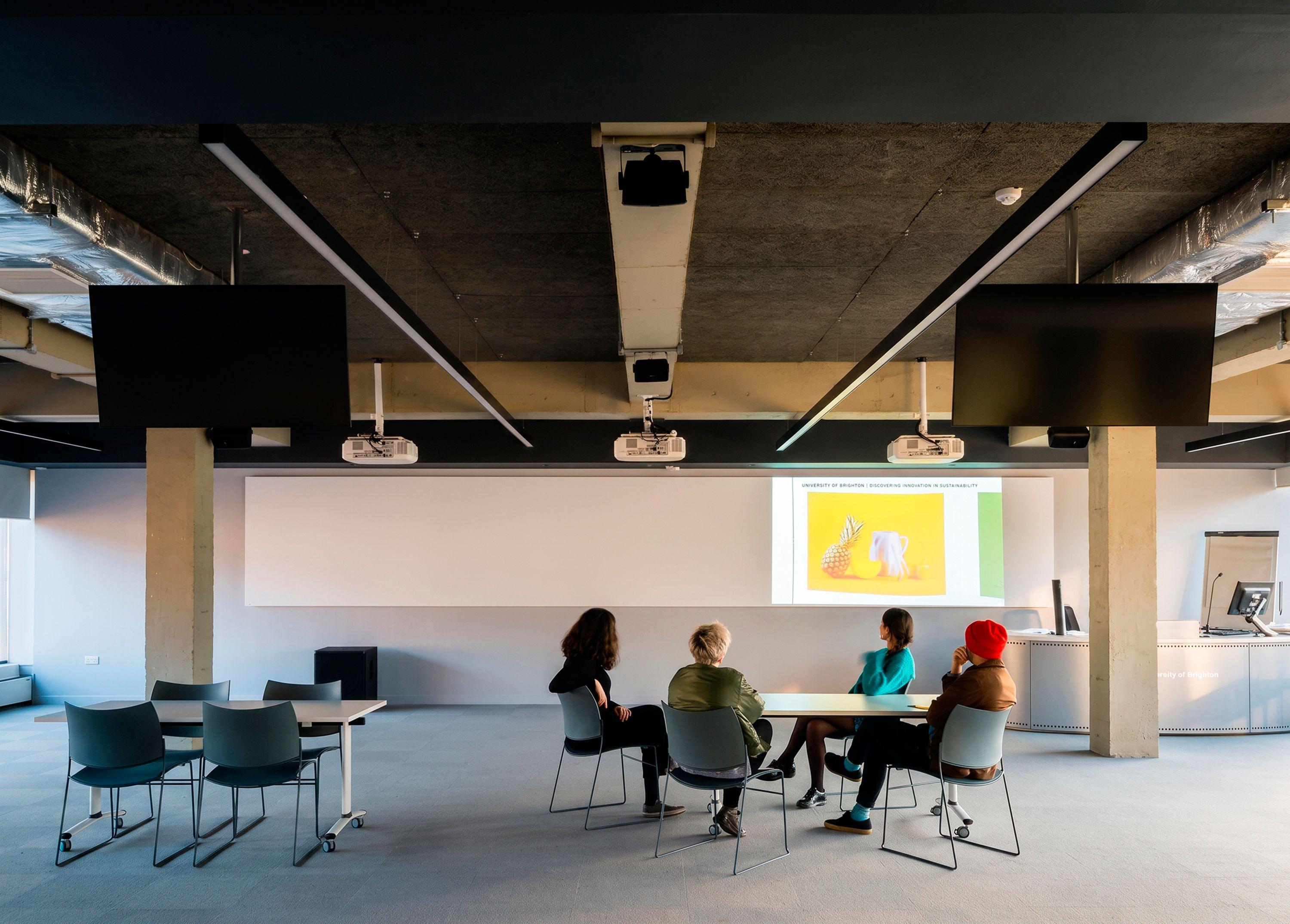 Formal teaching space at the University of Brighton photography and film centre, where students sit at a table facing a projected presentation, with the exposed concrete frame, ceiling-mounted projectors and screens overhead.