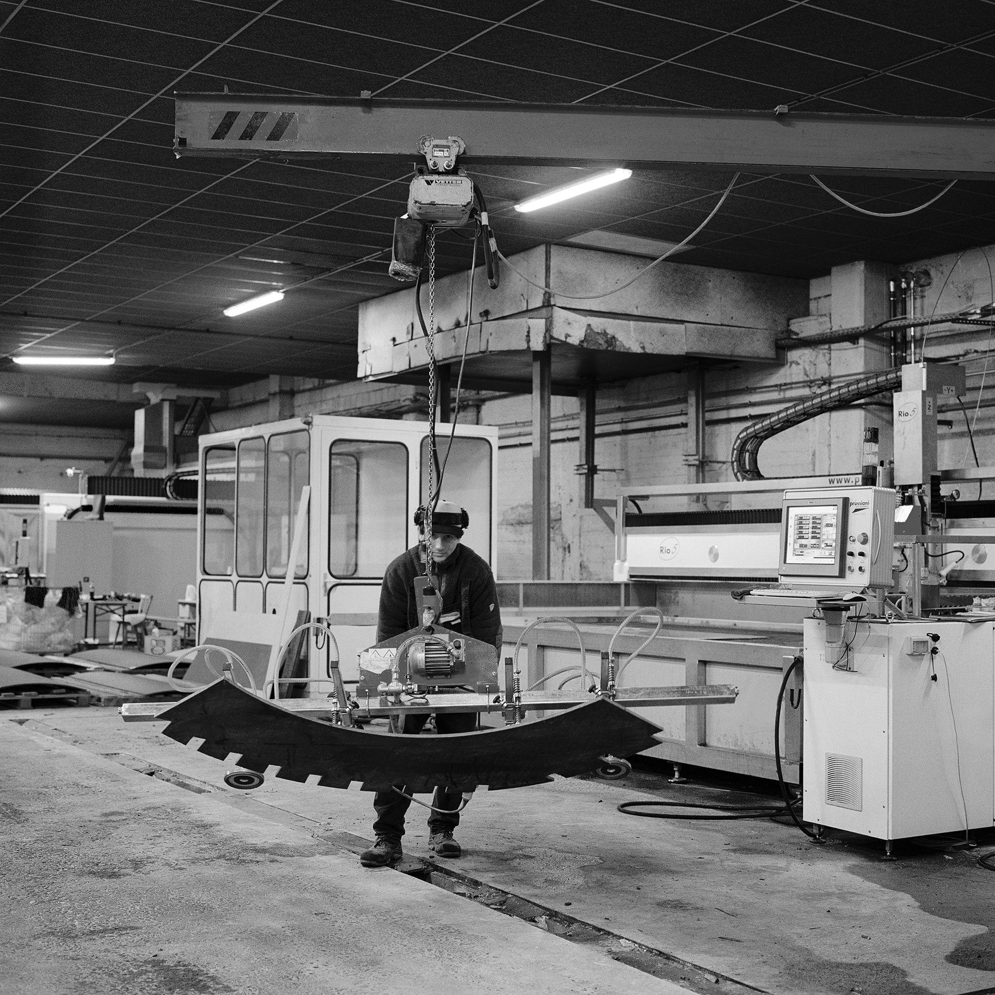 Curved concrete fin for the pavilion being lifted by chain hoist in a fabrication workshop, with cutting machinery behind.