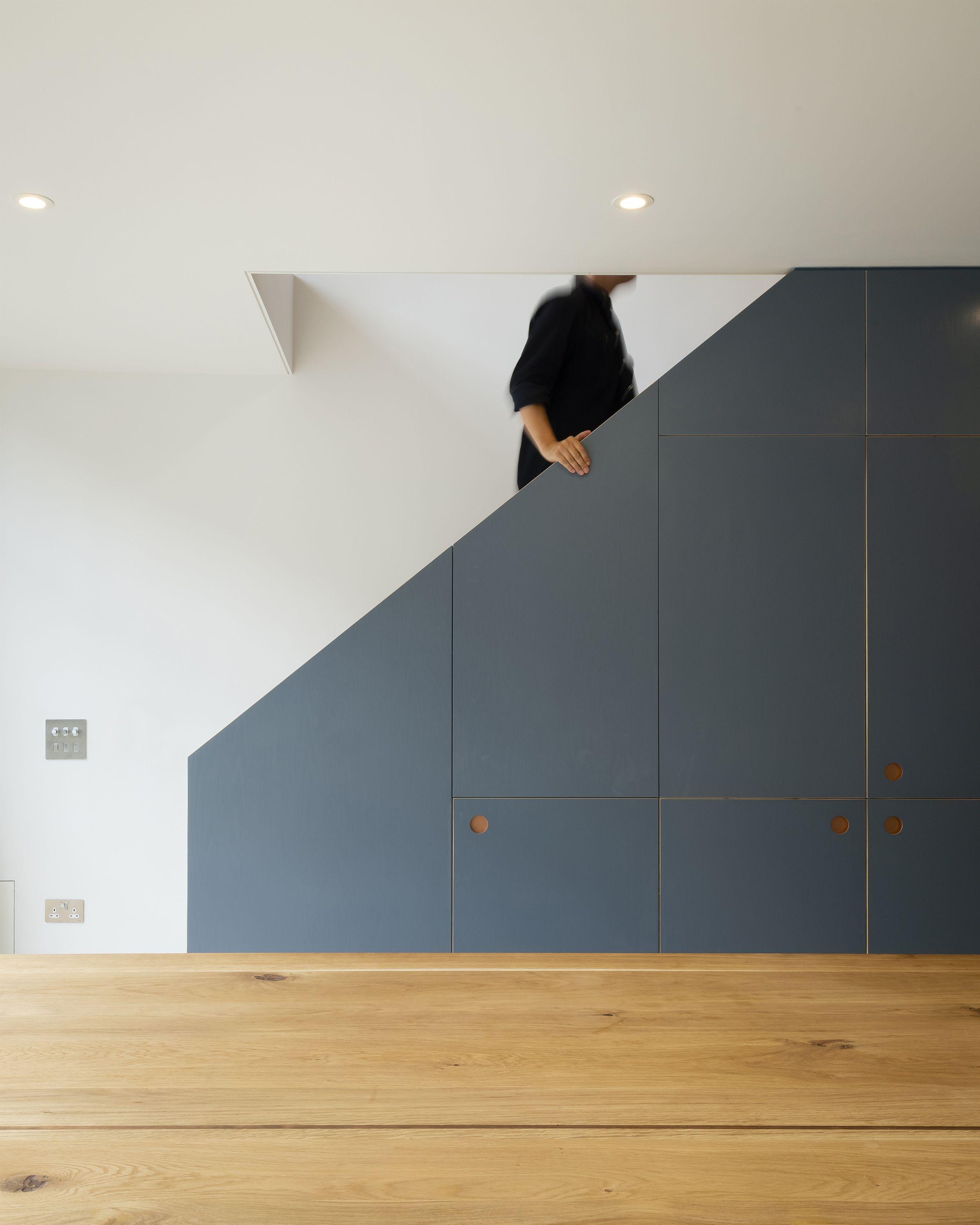 Grey-blue understair storage joinery at the Fluid Workspace, its stepped cupboard fronts with circular pulls set above a pale timber floor, a figure descending beyond.