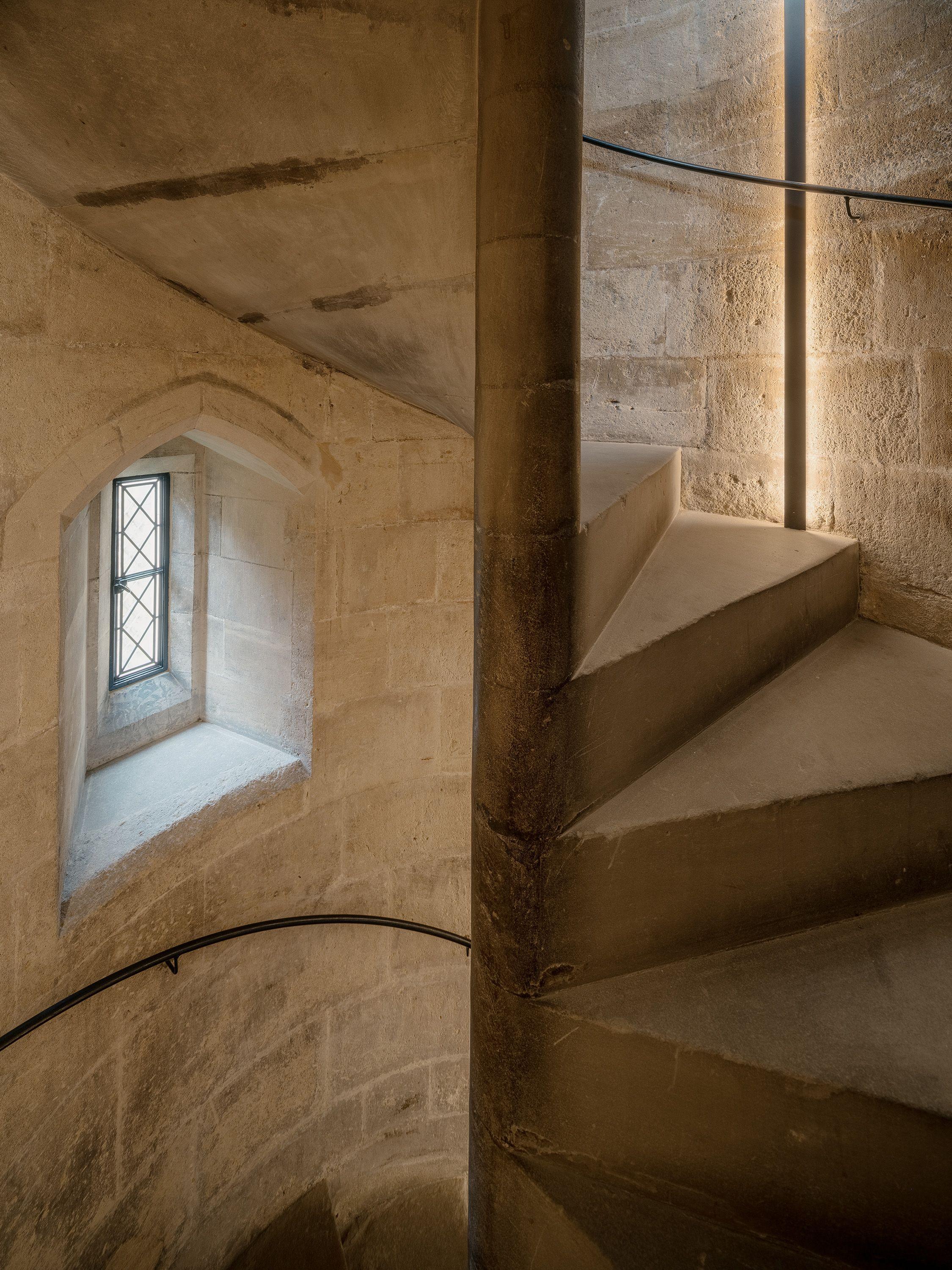 A narrow lighting slot set into the stone of the historic stair turret at Exeter College Library, Oxford, washing the ashlar and a single winding step in warm light.