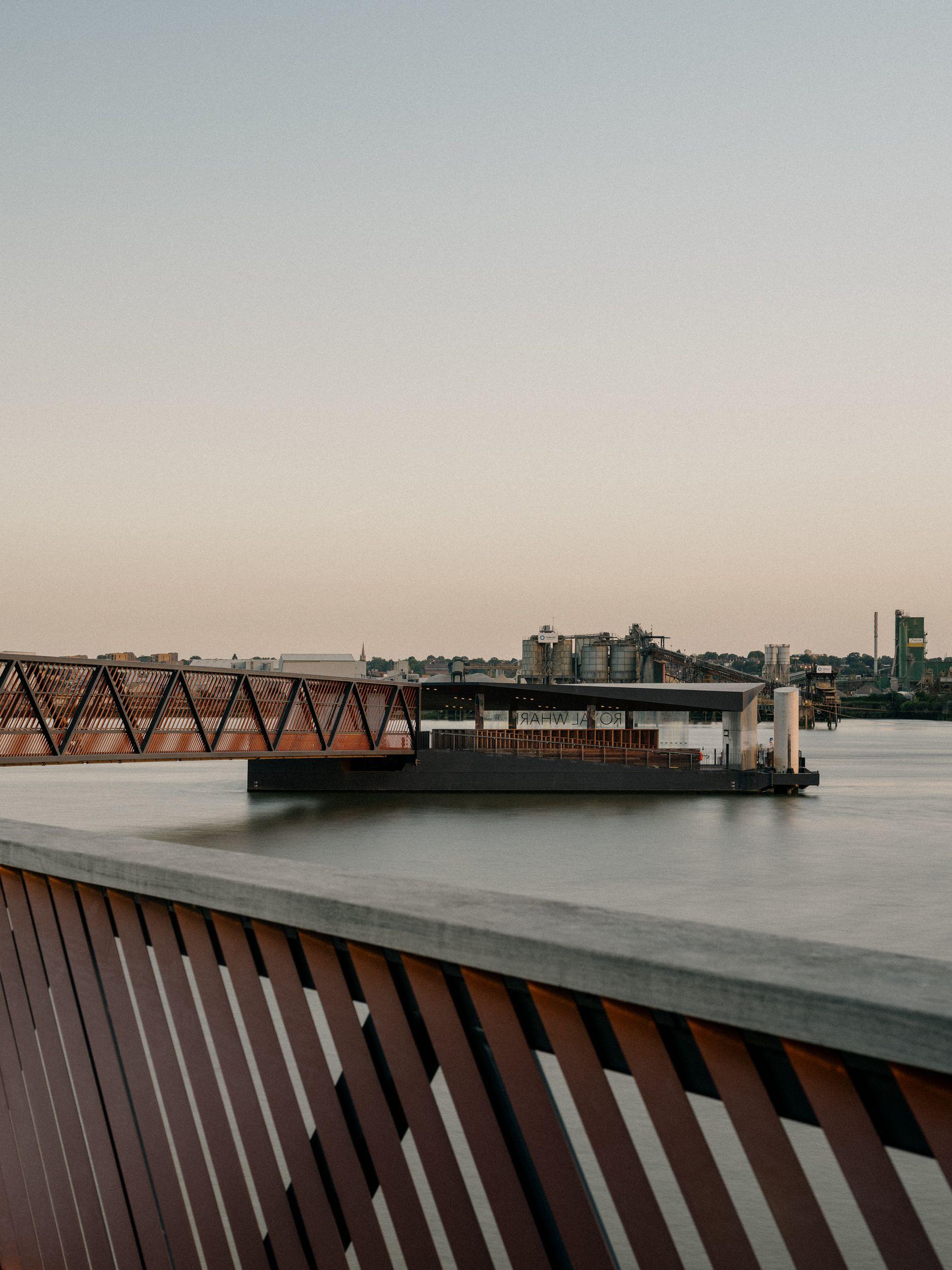 Royal Wharf Pier on the Thames at dusk, the timber and steel balustrade of the promenade running out towards the floating walkway and pontoon beyond.