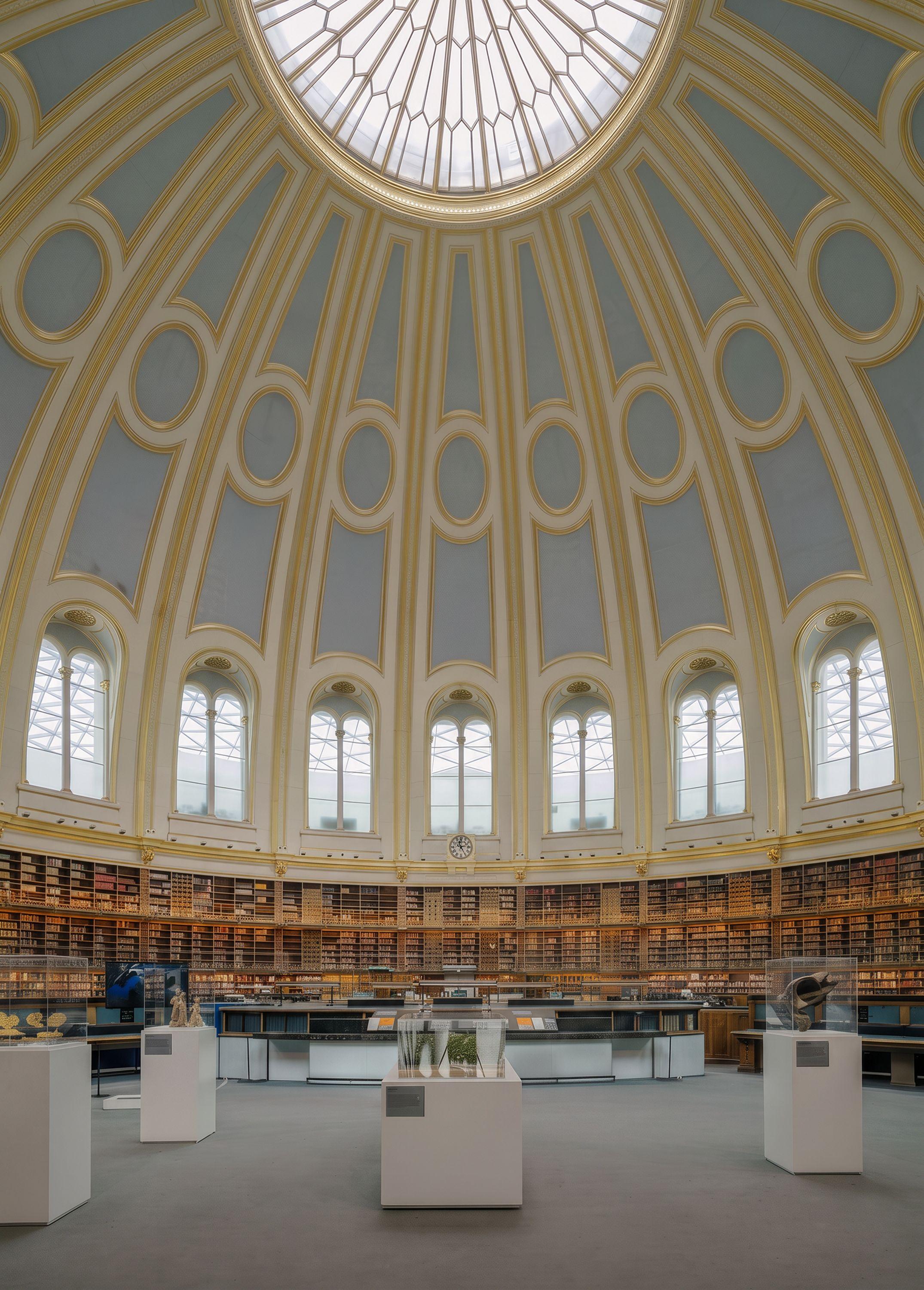 The domed reading room at the British Museum seen from the floor, gilded ribs and diffuse top light rising above the circular runs of bookshelves and windows.