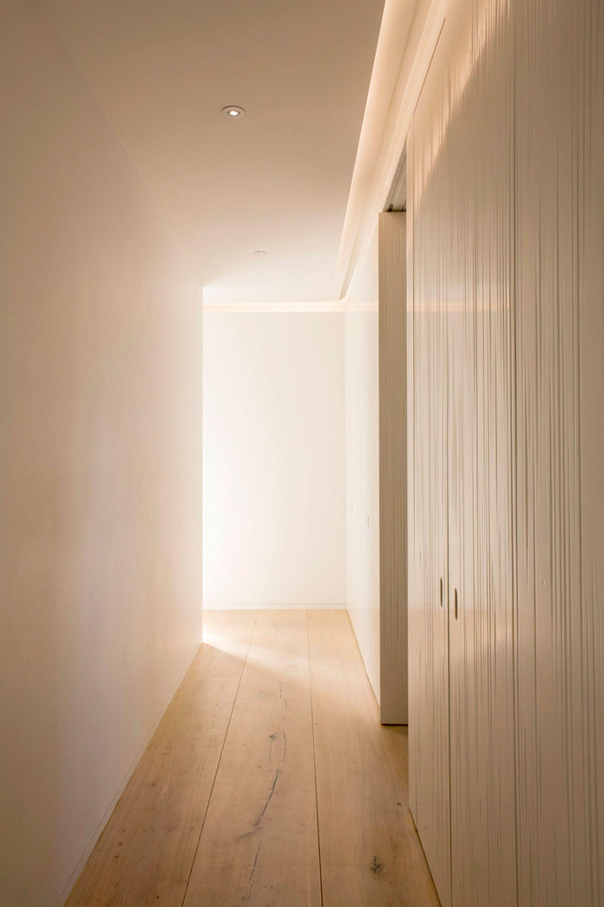 A hallway in a refurbished apartment, warm light falling across wide oak floorboards, a wall of textured timber joinery with concealed doors to one side