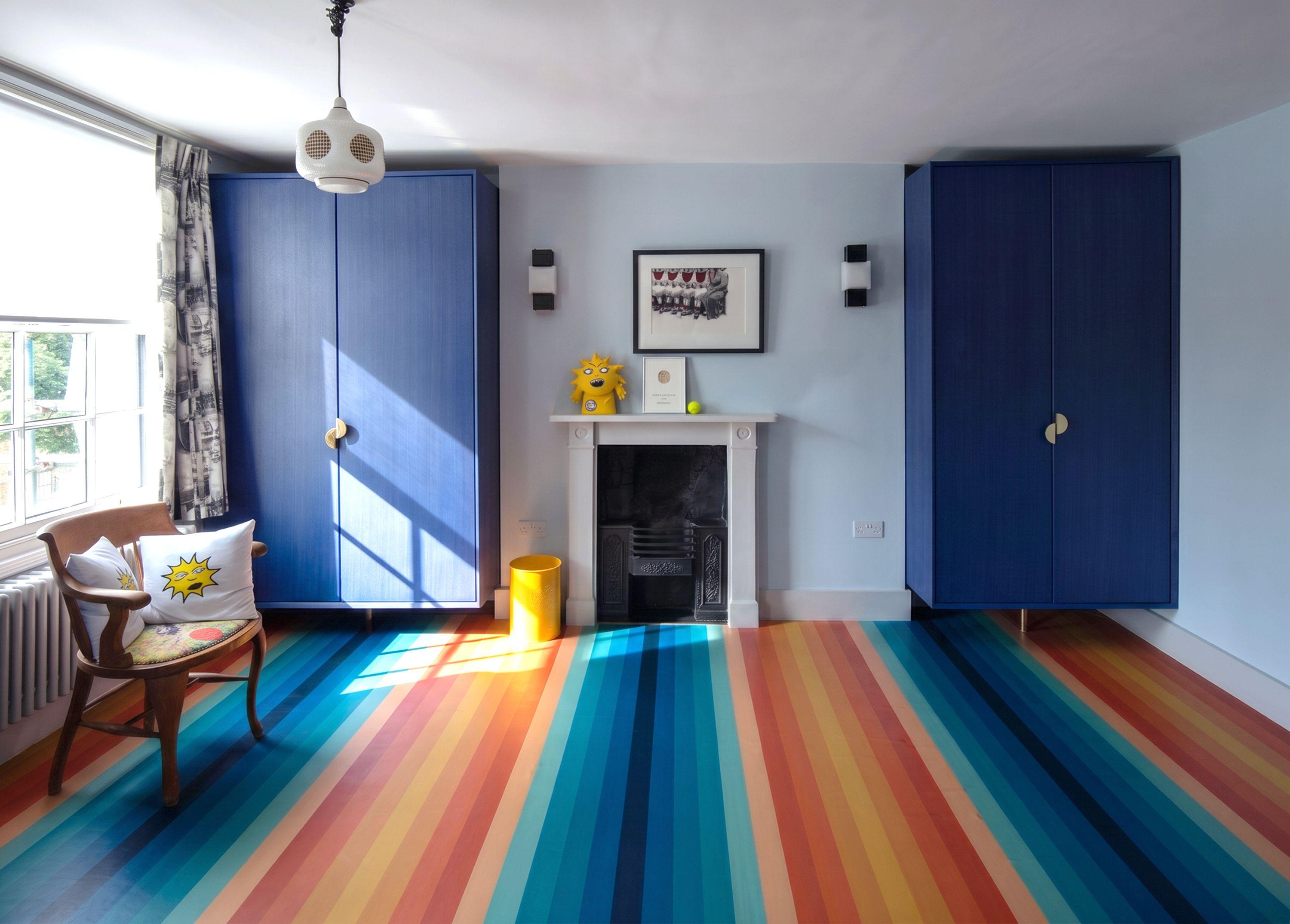 Child's bedroom at Puzzle Box with a striped rainbow floor, blue built-in wardrobes flanking a Victorian fireplace and pale blue walls.