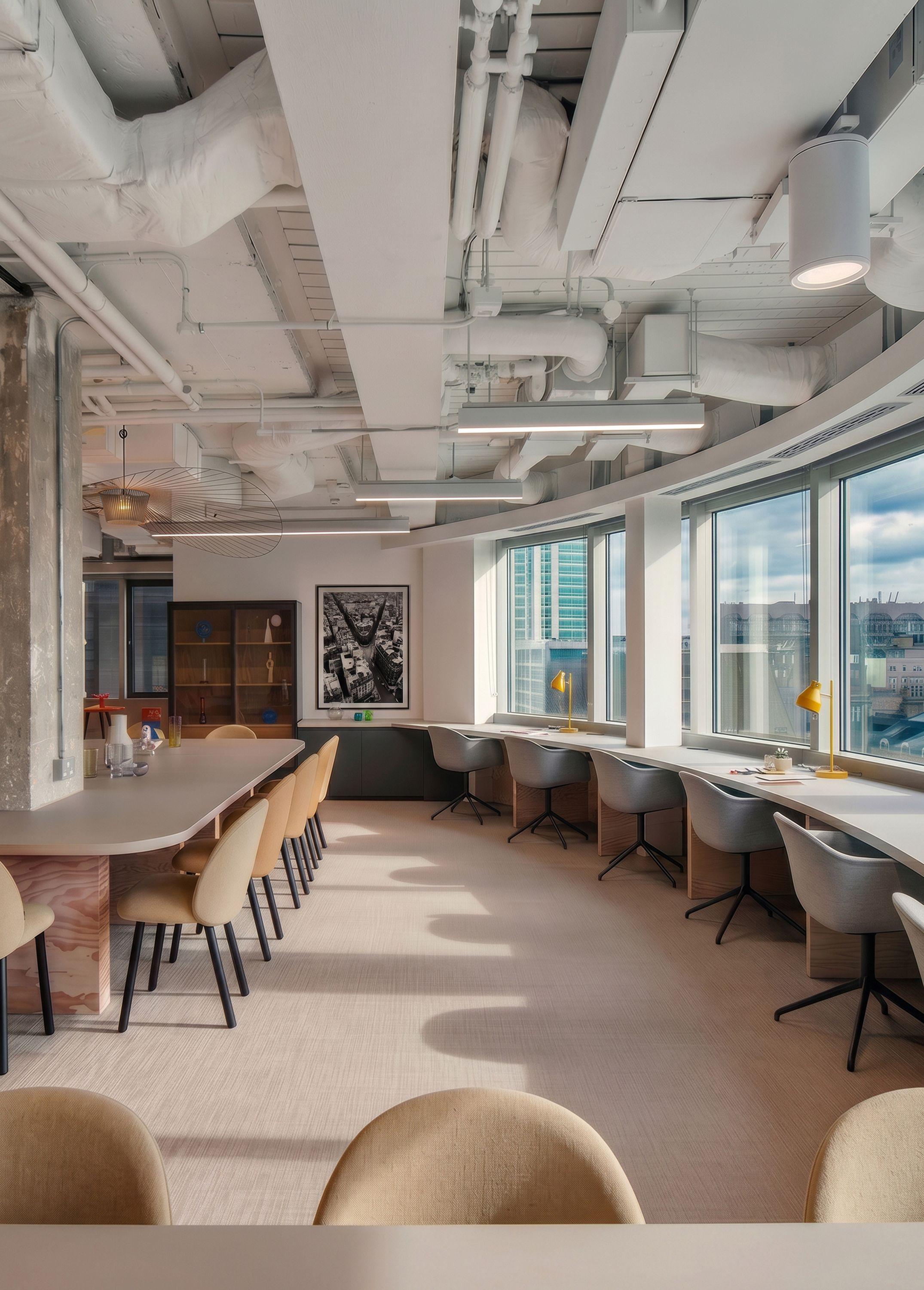 Communal dining area at Coppice Workplace, a run of tables set beside desk windows with the skyline beyond.