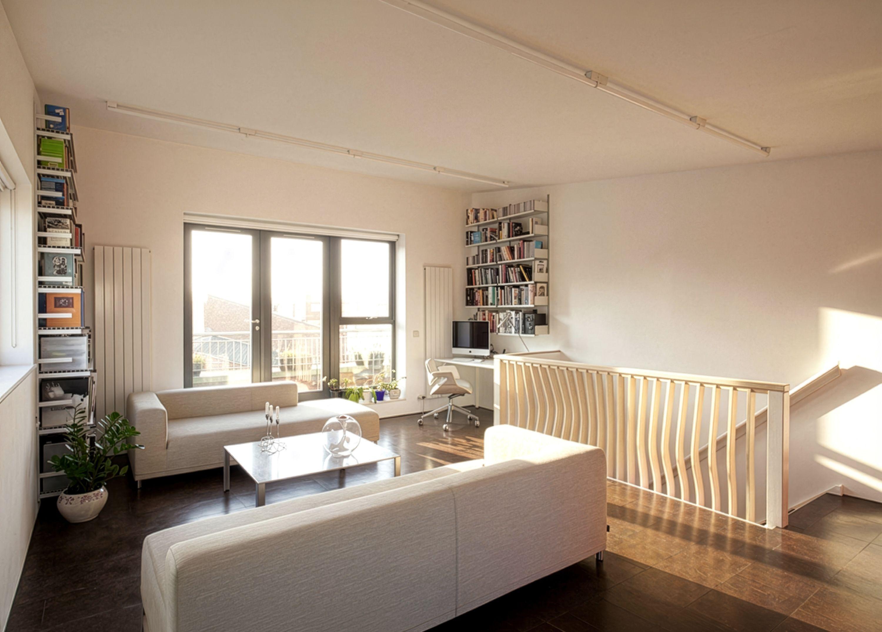 An open-plan apartment living room in warm light, the sculpted timber fin balustrade curving around the stair, sofas and bookshelves by the window